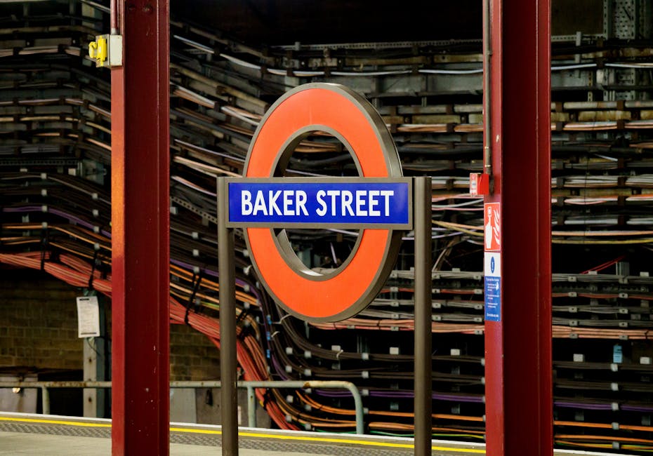 A close-up of a London Underground station sign reading 'Baker Street' in white text on a blue background, framed by a large, round, orange and grey circle. The background features a stack of black an