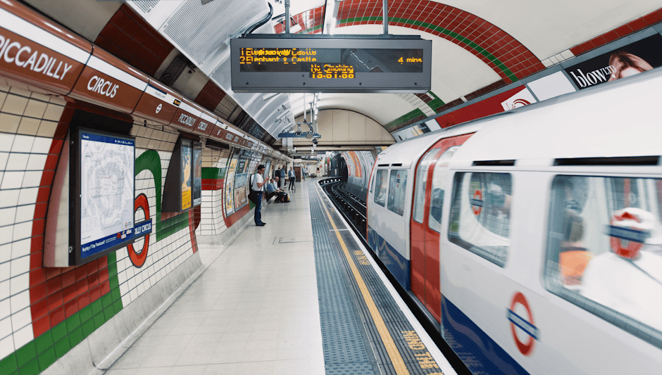 The London Underground platform at Earls Court station with a white and red train arriving, tiled walls in red, white, and green, and digital displays showing train information. There are a few passen