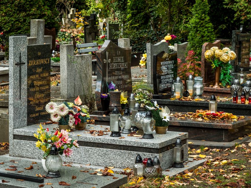 A collection of gravestones in a cemetery, decorated with an assortment of fresh floral arrangements and bouquets. The floral displays include white daisies with pink accents, pink lilies, and yellow