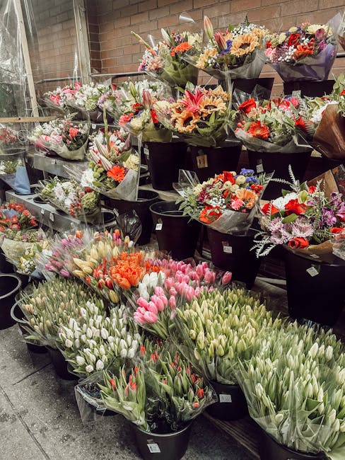 An outdoor display of numerous floral bouquets and bunches arranged in black buckets and wrapping containers. The bouquets feature a variety of fresh flowers including pink and white tulips, orange ro