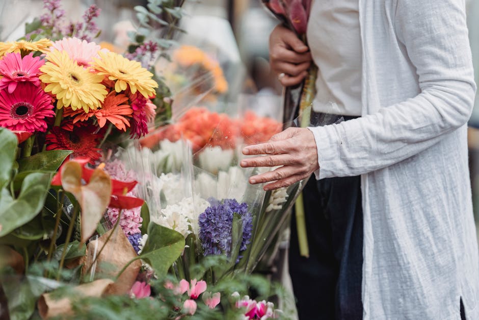 A person wearing a white long-sleeve shirt and dark trousers is examining a vibrant display of fresh flowers at a flower market. The arrangement includes bright pink, yellow, and orange gerbera daisie